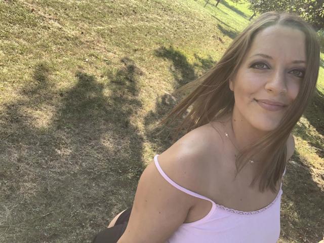 Lollipop69-hot with brown hair, pink top, and visible tits, smiling outdoors, sun casting shadows, grassy background, wearing a delicate necklace, looking at camera.