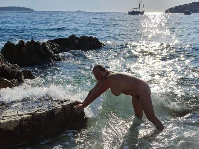 Naked Jutta1-hot with big boobs and ass, bending over rocky shore, water splashing, sun shining on wet skin, sailboat in background, blue ocean, rocks, clear sky.