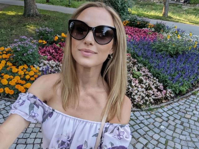 VickyFox, aantrekkelijk met blond haar, een paars bloemenblousje met open schouders, zwarte zonnebril en een subtiele glimlach, staat op een geplaveid pad, waarbij haar borsten en billen licht zichtbaar zijn..