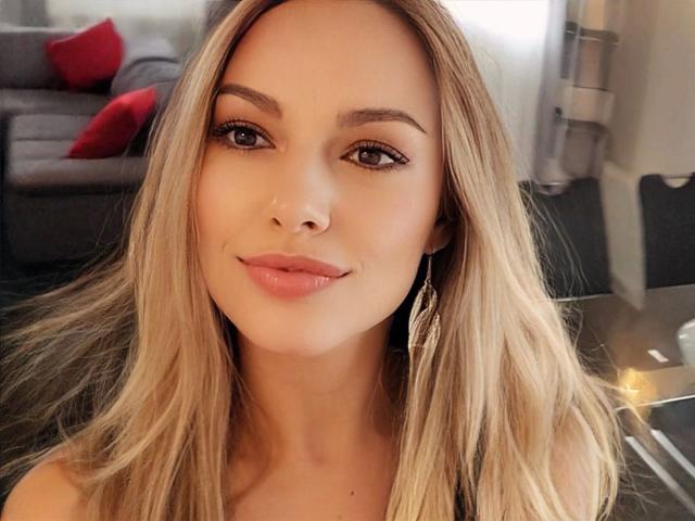VickyFox-hot heeft blond haar, bruine ogen en glanzende, roze lippen. Ze glimlacht zachtjes, draagt subtiele make-up en een zwarte top. Haar borsten zijn niet zichtbaar..