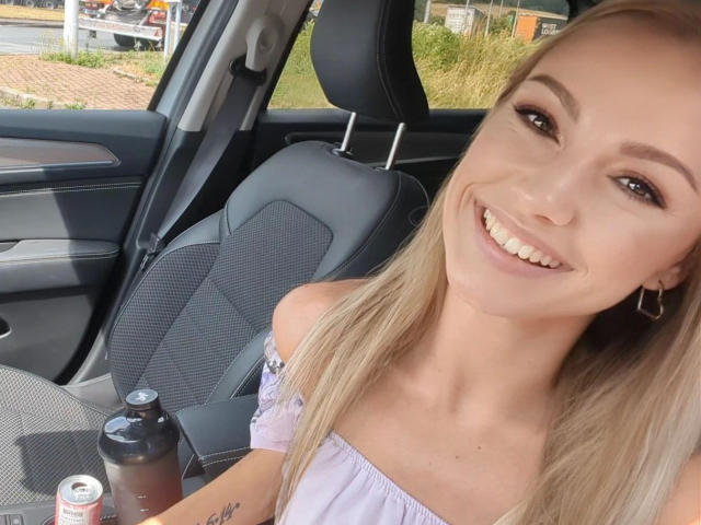 VickyFox, aantrekkelijk met blond haar, lachend, gekleed in een lila top met open schouder, zittend in een auto, met een blikje en een fles in haar handen, waarbij haar borsten en een kleine tatoeage zichtbaar zijn..