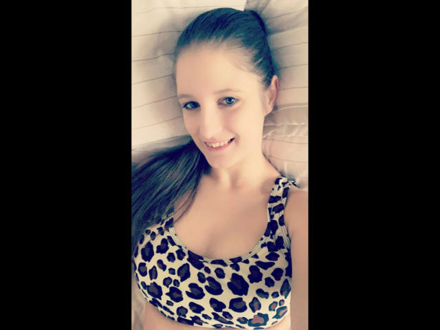 Jenniferamton-hot with long brown hair, smiling, wearing a leopard print sports bra, lying on a bed, showing her tits. She has light skin and blue eyes.