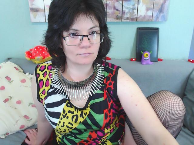 EverlyRays with short black hair, glasses, and a colorful leopard print dress, wearing a spiked necklace and fishnet stockings, sitting on a gray couch with a purple toy.