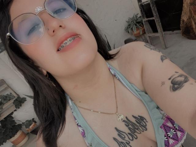 NatalieHarris with blue eyeshadow, glasses, black hair, tattoos, necklace, green halter top, showing teeth with metal braces, and a necklace with a pentagram. Her tits are visible.