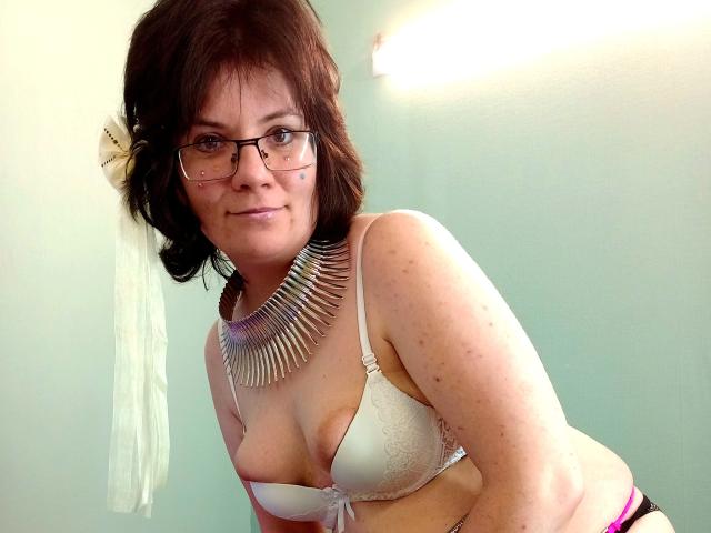 EverlyRays with brown hair, glasses, and a white lace bra, wearing a spiked necklace, leaning forward, showing her tits, and a pink thong, smiling.
