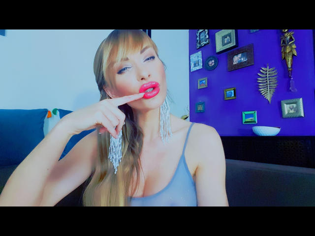 GiliSky-hot with red lips, finger in mouth, gray tank top, long blonde hair, white earrings, purple wall, brown couch, and various framed pictures in the background.