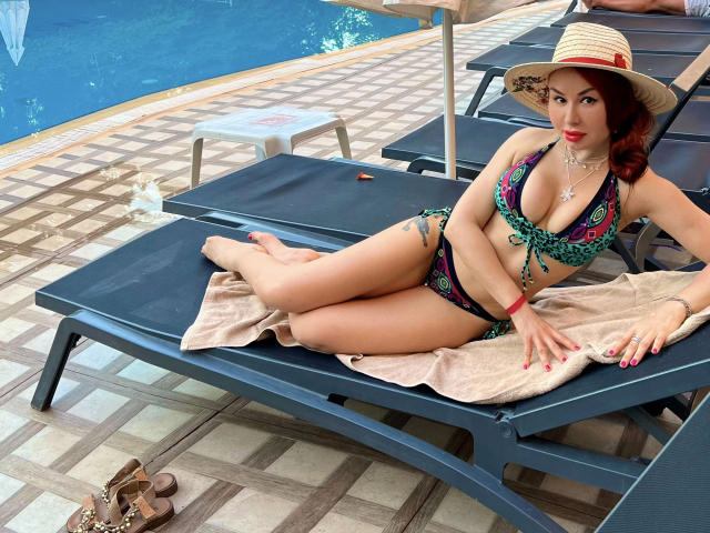 Catlady-hot with red hair, wearing a colorful bikini and straw hat, lounging on a blue pool chair, showing her tits, with tan towel and brown sandals on the tile.