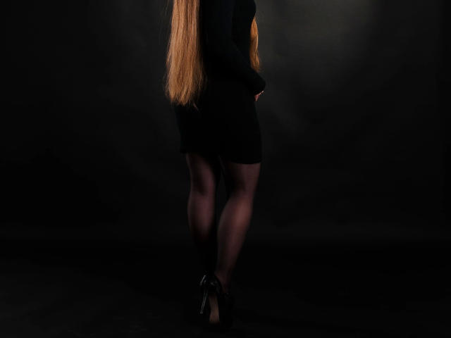 Junge Sekretärin – attraktiv in einem schwarzen Kleid und High Heels, langes blondes Haar, steht mit den Händen an den Seiten, Beine gekreuzt, dunkler Hintergrund, schwarze Strumpfhosen, schwarze High Heels, schlank und sexy..