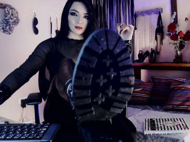 MisssDarkLady with pale skin, black hair, and red lipstick, showing the sole of her boot to the camera, wearing black gloves and sitting in a chair. Boobs partially visible.