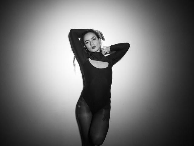 MaiaPark in a black, cut-out, long-sleeve leotard, standing with arms raised, showing her tits and ass, in a dark, gradient, high-contrast, black-and-white photo.