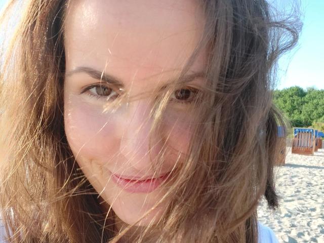 Schneewitchen1-hot has light brown hair, wind-blown face, and a slight smile with pink lips. Brown eyes are partially visible through tangled strands. She's wearing a white top.
