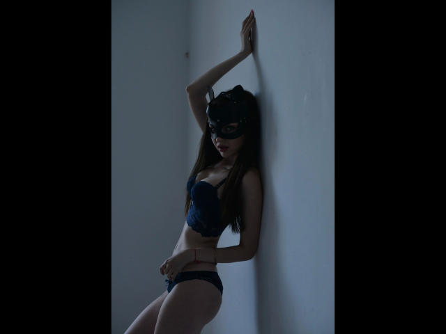 Lovly19, attraente, con lunghi capelli castani, indossa una maschera nera, un reggiseno di pizzo blu e mutandine abbinate, appoggiata a un muro bianco, mostrando il sedere e piccoli seni, in una luce soffusa..