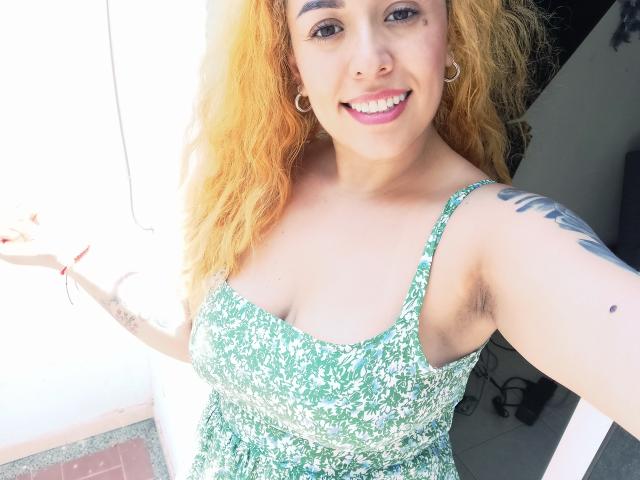 Joanallen with orange curly hair, green floral dress, showing armpit hair, small tattoo on right arm, smiling, hoop earrings, pink lipstick, and a cute nose ring.