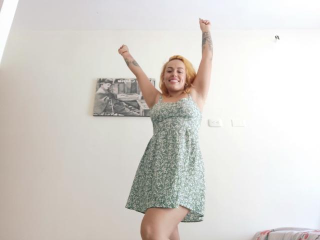 Joanallen has blonde hair, smiling, arms raised, wearing a green floral dress, tattoos on her arms, standing in a bright room, smiling with joy.