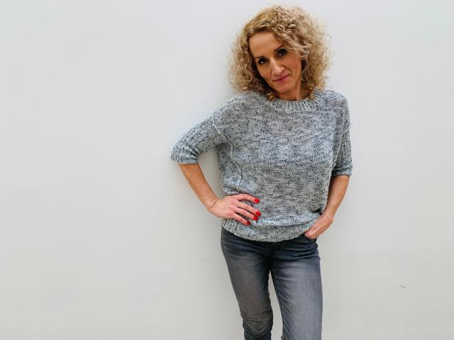 SusaneNori-hot with curly blonde hair, gray knit sweater, blue jeans, red nails, hands on hips, smiling slightly, standing against a white wall.
