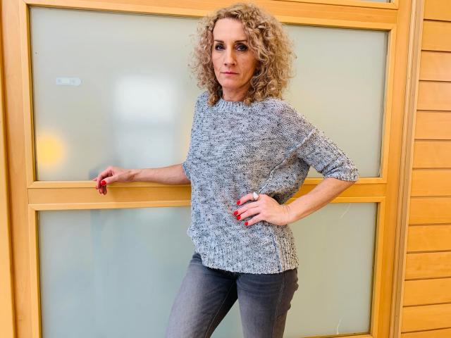 SusaneNori-hot with curly blonde hair, gray knit sweater, and blue jeans, standing with hand on hip and other arm on wooden door, red nails, confident smirk.