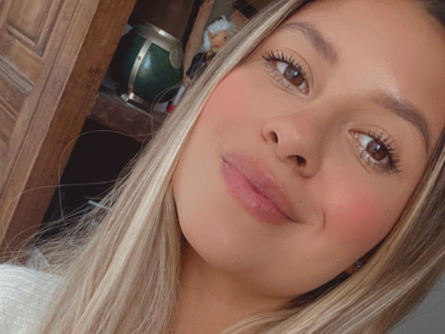 AniCuteLove's face with blonde hair, brown eyes, and pinkish lips, wearing a white top, showing a subtle smile. Her ass is not visible. Her tits are not visible.