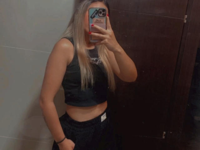AniCuteLove with blonde hair, black crop top, and black pants, holding phone to cover face, showing her bare midriff and small ass, in a mirror selfie.