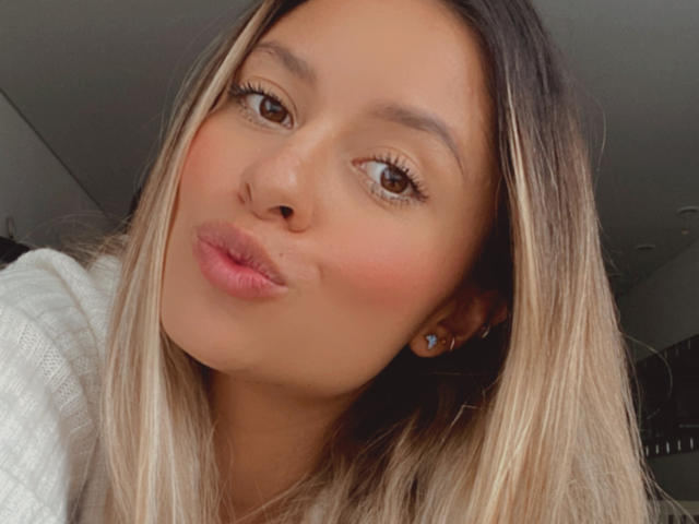 AniCuteLove's face, showing brown eyes, blonde hair with dark roots, pinkish lipstick, slight pout, white sweater, small blue earrings, light brown skin, soft makeup, slightly tilted head.