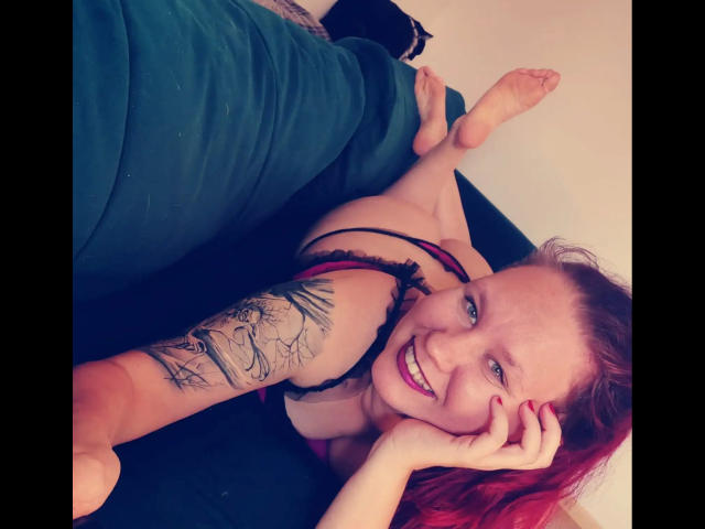 Suniabdsm-hot with red hair, smiling, lying on a green couch, showing tattooed arm, wearing black and pink lingerie, legs up, bare feet, red nail polish, cute, sexy.