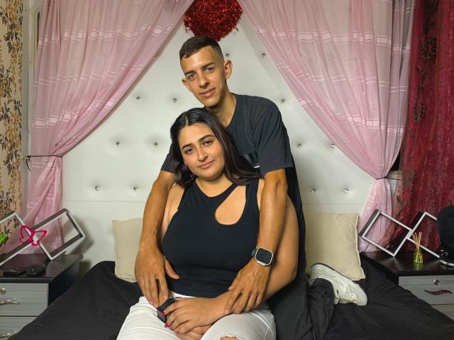 KattaAndJhon, avec une peau marron clair, un débardeur noir, un pantalon blanc, assis sur un lit. Jhon, avec des cheveux bruns courts, une chemise noire, une montre, les bras autour des épaules de KattaAndJhon..