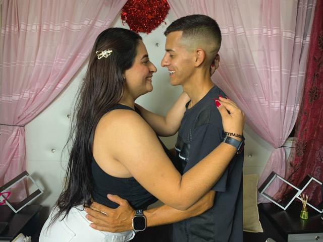 KattaAndJhon, with long dark hair and big boobs in a black tank top, hugs Jhon, who's smiling with short brown hair, wearing a gray shirt, in a room.