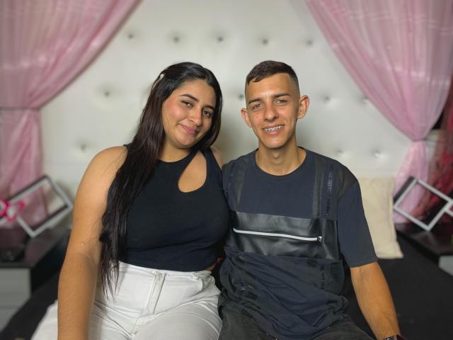 KattaAndJhon, both sitting on a bed. Katta has long black hair, white pants, black top, big tits, and a cute smile. Jhon has short brown hair, black shirt, and black straps.
