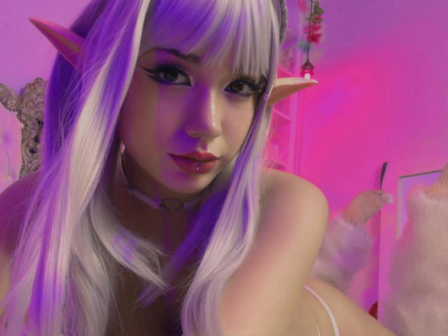 SabriPrettty with long white hair, elf ears, and purple makeup, has puffy pink lips, and is wearing a choker, looking at the camera with a sexy expression.