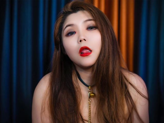 Laruina-hot with long brown hair, red lipstick, and a black collar with a gold bell, sticking her tongue out, staring with blue eyes, showing her bare shoulders.