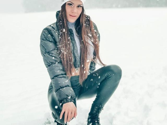 Mia124-hot, squatting in snow, wearing a grey puffy jacket, black leather pants, and black boots, with long brown hair, and a white beanie, snowflakes on her face.