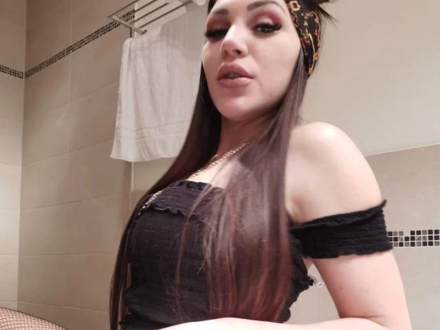 Mia124-hot with long brown hair, wearing a black off-shoulder top, leopard headband, and gold necklace, looking at the camera with pouty lips, in a bathroom.