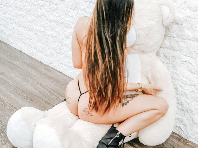 Mia124-hot, topless, with long brown hair, sitting on a giant white teddy bear, wearing black thong and boots, showing ass and tattoo on right thigh.