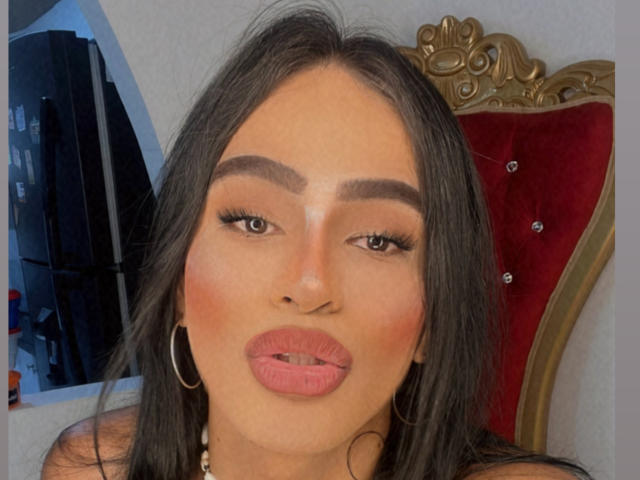 IrinaFerrero's face, with full pink lips slightly parted, dark eyebrows, long black hair, and gold hoop earrings, sits in a red velvet chair with a gold ornate back.