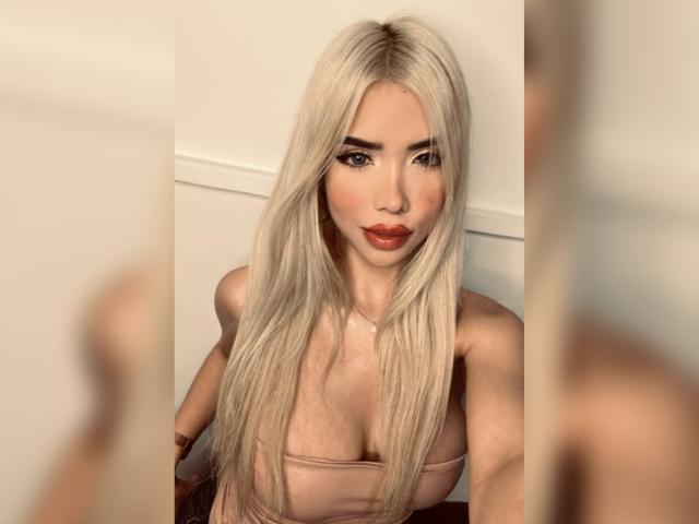ChristieGarci with long, platinum blonde hair, red lipstick, and a pink top, showing her tits in a selfie. Her eyes are blue, and she's looking at the camera.