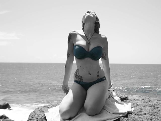 BeachSchnecke, in ginocchio su delle rocce, indossa un bikini verde, la testa è inclinata all'indietro, ha tatuaggi sulle braccia e sul fianco, le mani sono sulle cosce, l'oceano sullo sfondo, foto in bianco e nero con bikini verde..