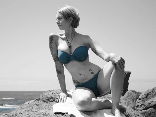 BeachSchnecke-une femme avec des cheveux blonds courts, un bikini bleu, des tatouages, assise sur des rochers, le genou droit levé, le bras gauche reposant, souriante, photo en noir et blanc, rocher texturé, océan en arrière-plan..