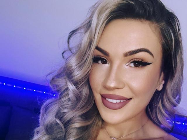 ZoeyMarie has blonde, wavy hair, full eyebrows, winged eyeliner, and dark pink lipstick, smiling at the camera with a cute, sexy face. Her tits are not visible.