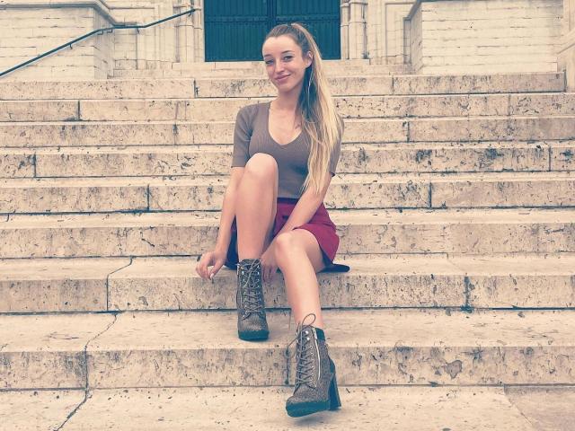 CremeDeLaCremeDE sentada en escalones de piedra, vistiendo una blusa marrón, una falda roja y botas de combate grises, sonriendo con las piernas cruzadas, con una larga coleta rubia y un rostro lindo..
