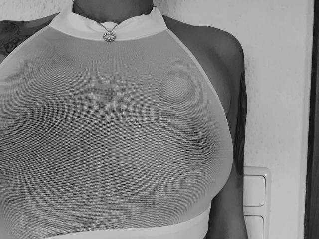AbenteuerMelody-hot's large, visible tits through a sheer, halter-top. The fabric's transparency shows dark nipples and a small, round belly button. Black and white, tight shot.