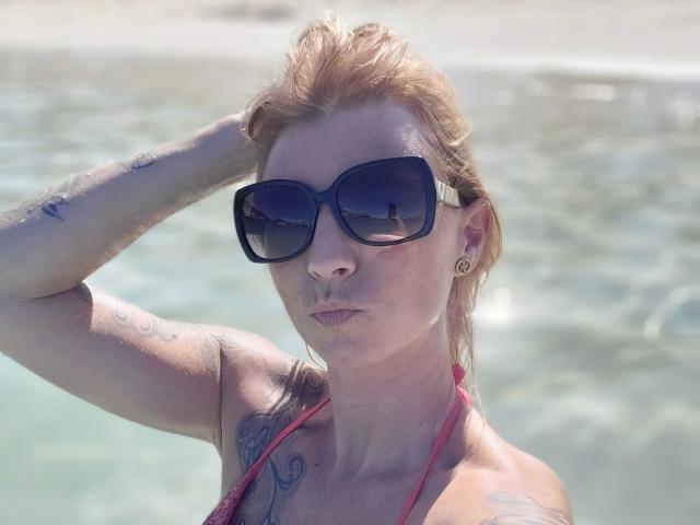AbenteuerMelody-hot with wet red hair, wearing big black sunglasses, red halter top, tattoos, and earrings, has a hand in her hair, and a pouty face by the water.