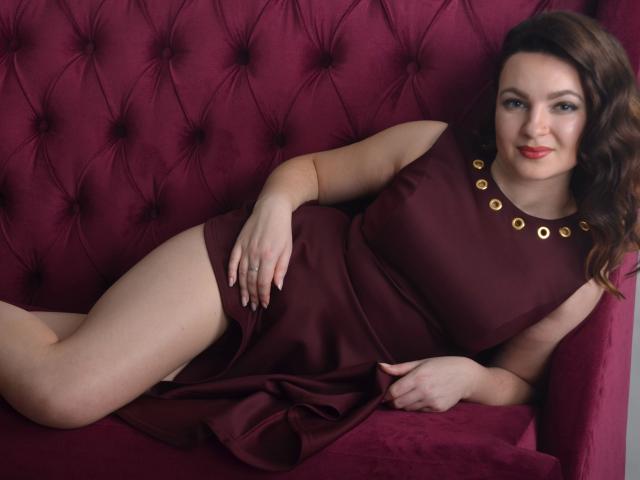 CaiseyMiss, relaxada em um sofá de veludo vermelho, vestindo um vestido marrom sem mangas com botões dourados, exibindo suas nádegas e coxas, com um sorriso tranquilo e cabelos castanhos ondulados..