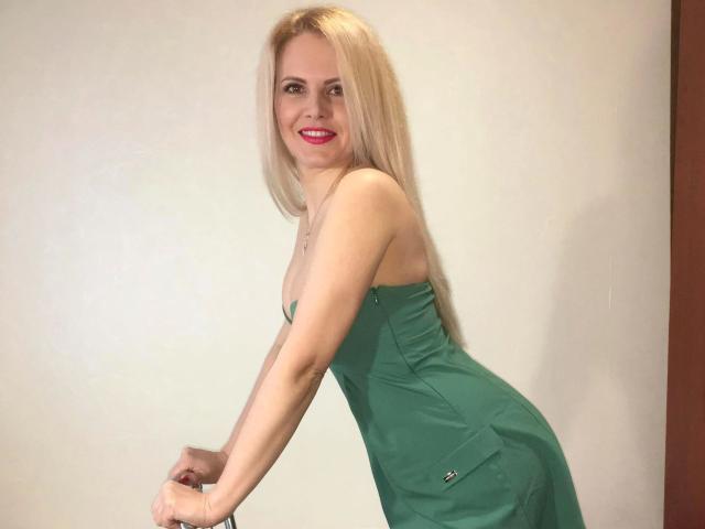 ReifeJuliette-hot with long blonde hair, green dress, and red lipstick, leaning forward, smiling, showing her ass and small tits, against a plain wall.