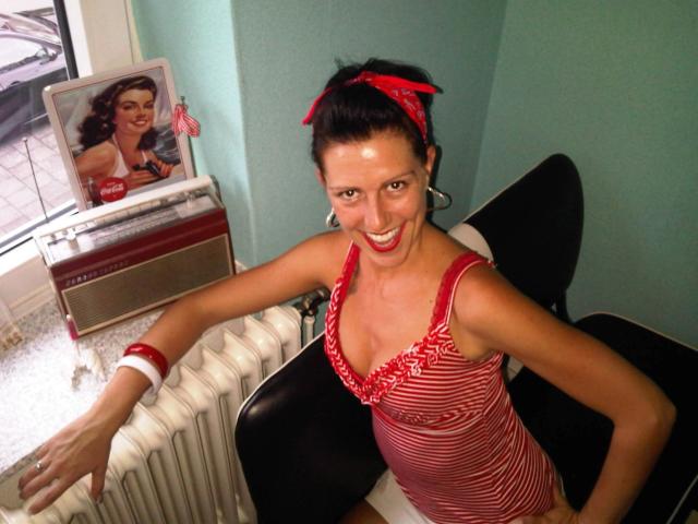 Texaspatti, souriante et avec du rouge à lèvres, portant un débardeur rayé rouge et blanc, montrant ses petites seins, un bandana rouge et un short blanc, appuyée sur une chaise noire..