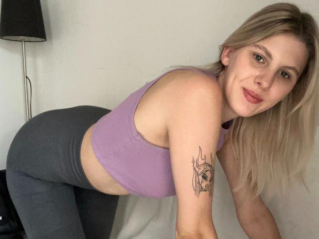 NicoleCherry-hot with blonde hair, purple sports bra, and gray pants, leaning forward, showing her ass, with a tattoo on her right arm, smiling at the camera.
