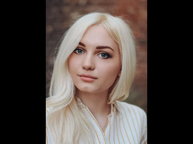 EvaTesla, con i suoi lunghi capelli biondo platino, gli occhi azzurri e un'espressione seria, indossa una camicia a righe bianche e gialle, ha un viso carino e labbra piene..