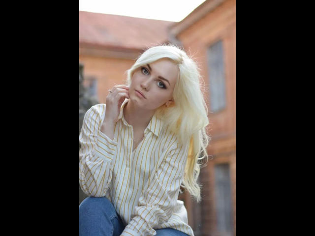 EvaTesla, met blond haar, een gestreepte blouse en blauwe jeans, in een hurk, met haar hand dicht bij haar kin, naar boven kijkend, lichtjes fronsend, op een onscherpe stedelijke achtergrond..