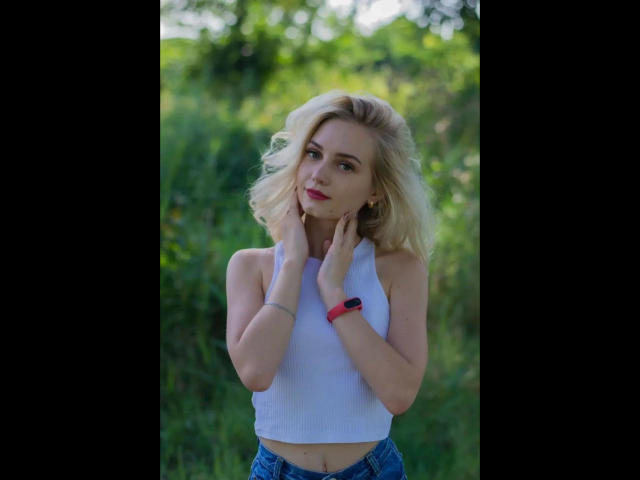 EvaTesla-hot heeft blond haar, draagt een witte mouwloze top en blauwe jeans, met haar handen op haar nek, en draagt een roze polshorloge, staand in het groene gras..