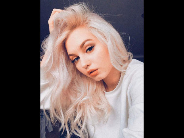 EvaTesla, con i capelli biondo platino e un aspetto sensuale, indossa un maglione bianco, guarda in basso con una mano tra i capelli, mostrando il seno e un'espressione accigliata..