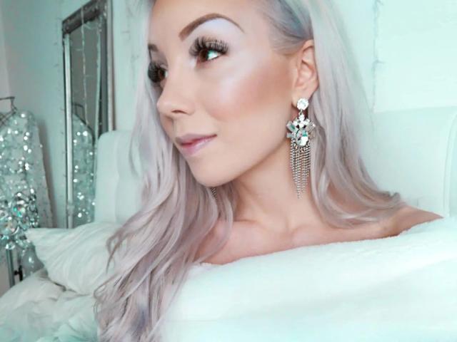 Lady-Khaleesi with long, platinum blonde hair, wearing dangly earrings, smiling, showing off her boobs in a white, off-shoulder top. She's got sexy, dark eyebrows and a cute nose.