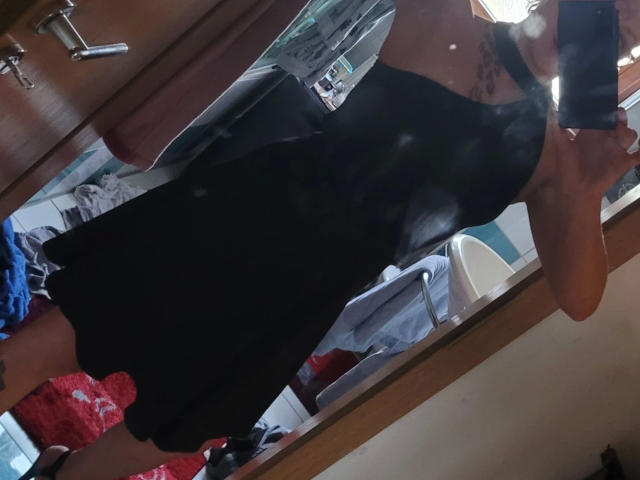 Mary-Licious in a black dress, showing her ass, holding a phone in front of a mirror. Visible tattoo on her left shoulder. Reflection includes her arm and part of her face.